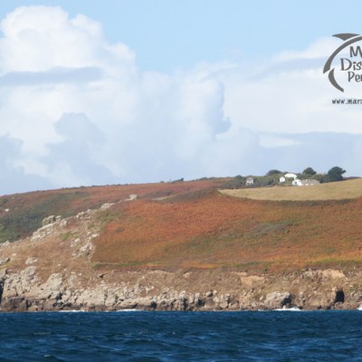 Cliffs with autumn foliage above a blue ocean, small buildings on top, cloudy sky.