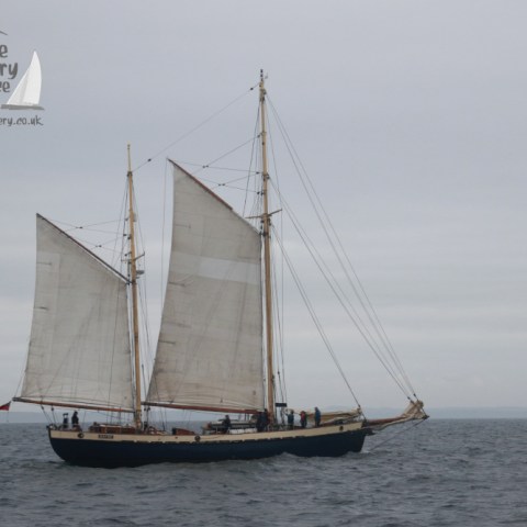 Sailing ship with two masts on open water, overcast sky, logo in the corner.
