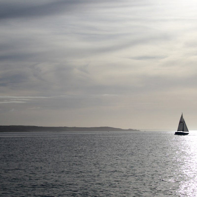 Sailboat on calm sea with distant ship and cloudy sky at sunset.
