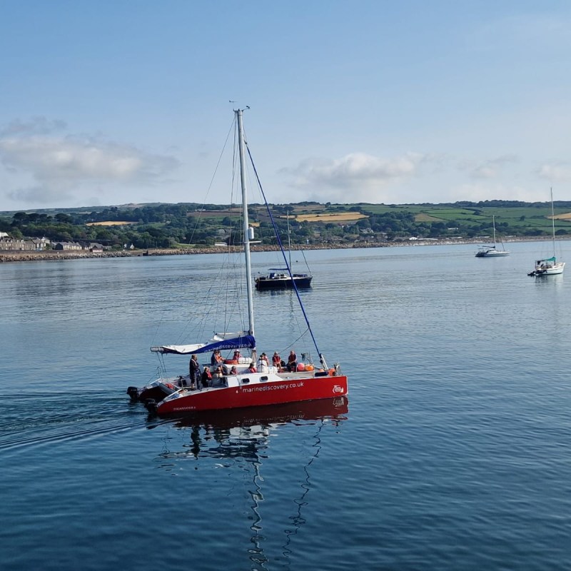 Red sailboat with people on a calm sea, with distant land and other boats.