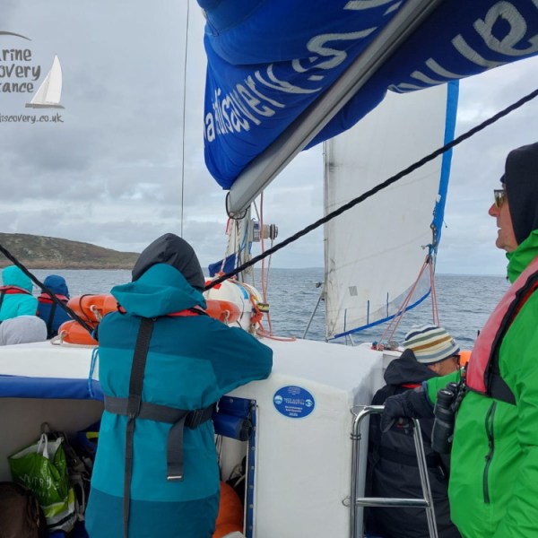 People in jackets on a sailboat, with a view of the sea and distant hills.