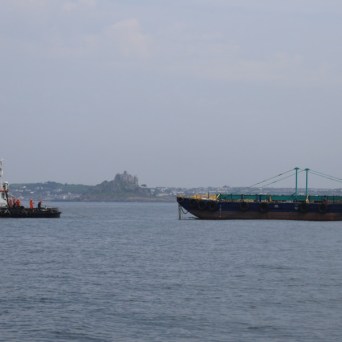 Tugboat towing a large barge in a calm sea, with distant coastline in the background.