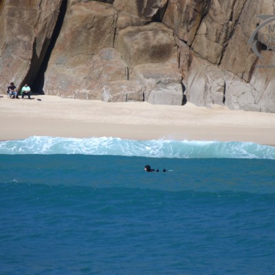 People on a beach with rocky cliffs, swimmer in the sea, and waves crashing on the shore.