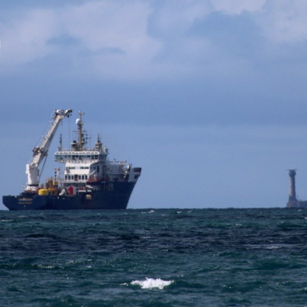 Large ship with crane at sea near rocky formations under cloudy sky.