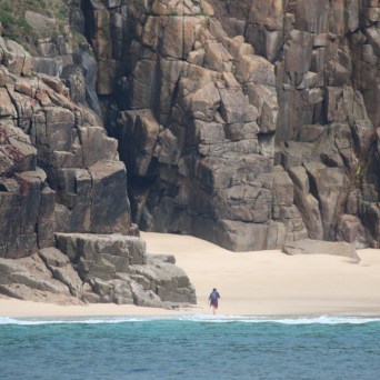 Person walking on a sandy beach with tall rocky cliffs in the background.