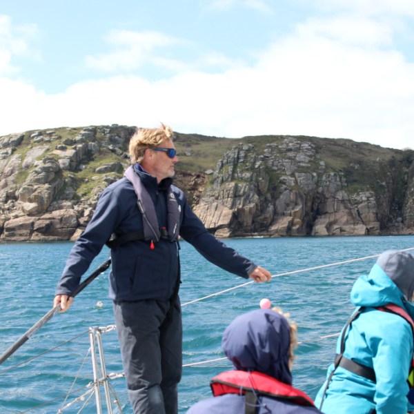 Group on a sailboat in front of rocky cliffs, person standing at the helm.