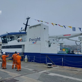 Three people in orange overalls by a docked freight ship with cranes.