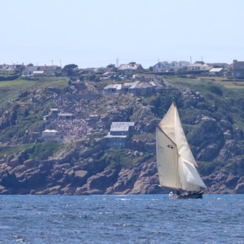 Sailboat on the ocean with rocky cliff and houses in background.