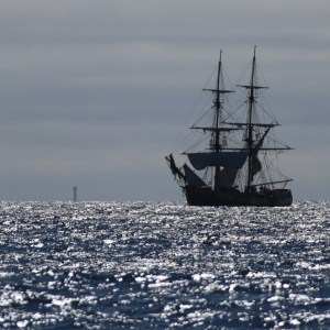 Silhouette of a large sailing ship on a sparkling sea under a cloudy sky.