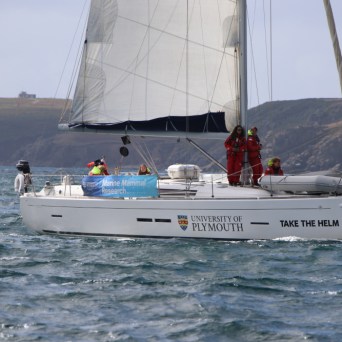Sailboat on ocean with people, banner says 'University of Plymouth', coastline in background.