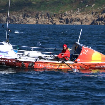 Small boat with colorful text in the ocean, person in red gear onboard, rocky shoreline in background.