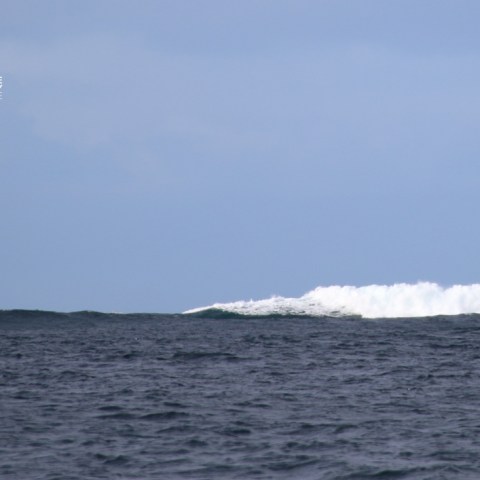 Boat on ocean near large breaking wave under blue sky.