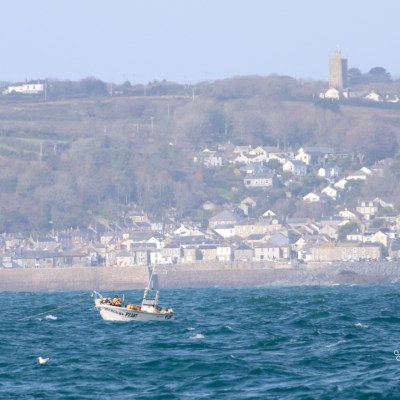 Small boat on ocean with coastal town and hills in background.